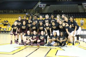 Women basketball players from both Appalachian State and UT Arlington pose for a picture at half court. They are all wearing t-shirts in support of Coach Angel Elderkin who was diagnosed with cancer.