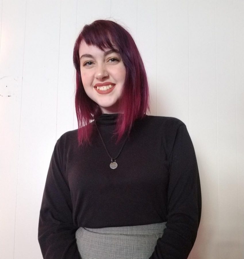 This is an image of Sophia, the chief copy editor. Sophia is wearing a black turtleneck shirt and high-waisted grey skirt. Sophia also has magenta hair and is smiling in front of a white background.