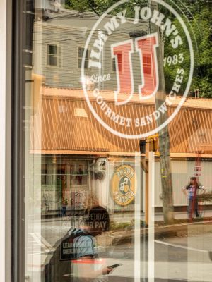 Boone Bagelry reflected in Jimmy Johns window across King Street Boone, NC. Hanging in the store is a sign that says Were competitive so is our pay. Boone Bagelry has been locally owned and operated since 1988.