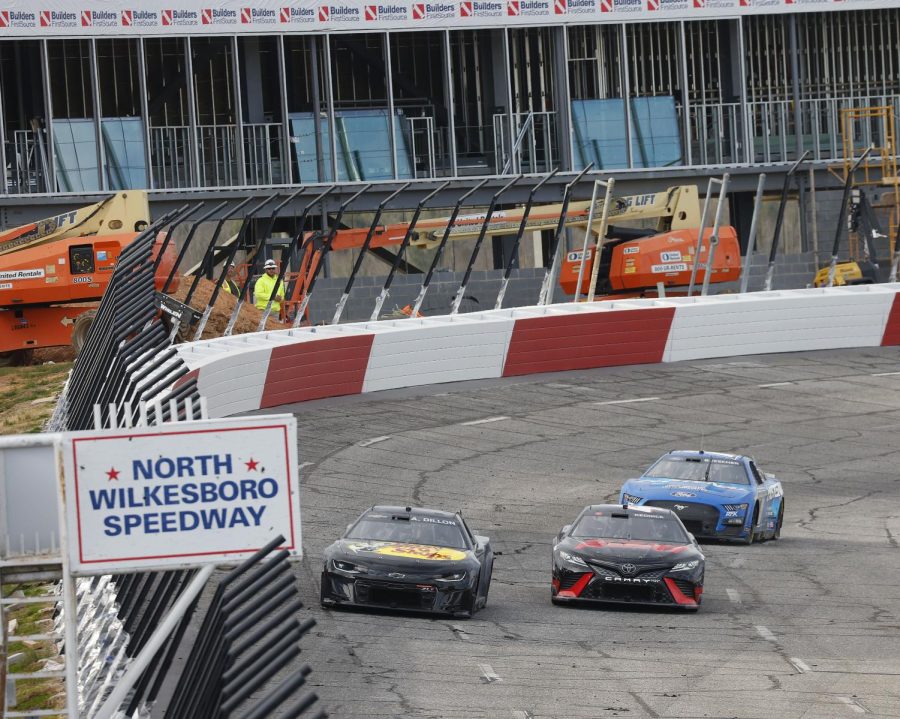 Three cars drive the North Wilkesboro Speedway as construction takes place outside the track.