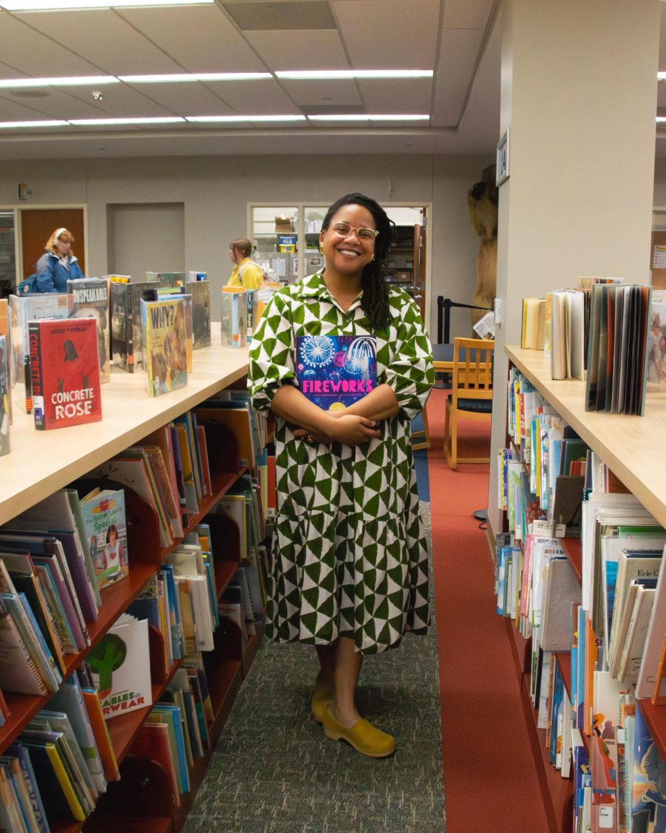Jewel Davis standing in the bottom floor of the library around the children's books. She holds this year's winning books Fireworks by author Matthew Burgess and illustrator Cátia Chien.